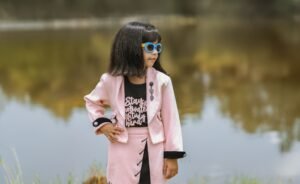 A fashionable child wearing sunglasses and a pink outfit poses by a serene lake.