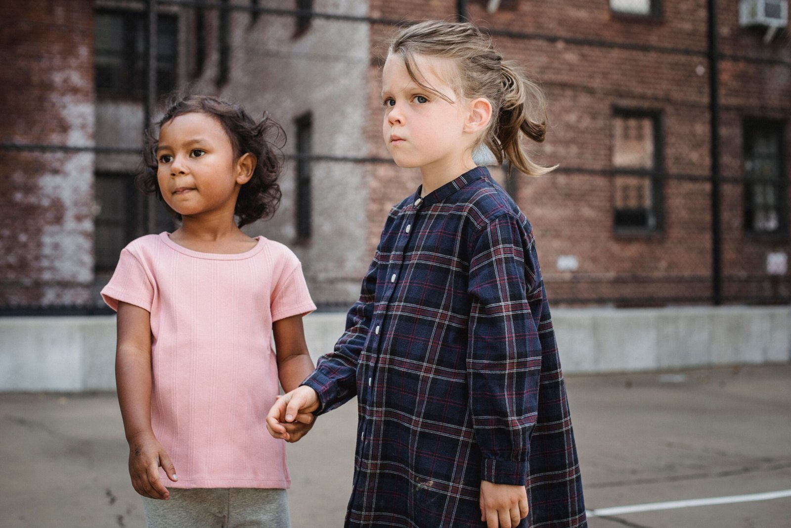 Two young girls holding hands, standing in an urban playground setting.