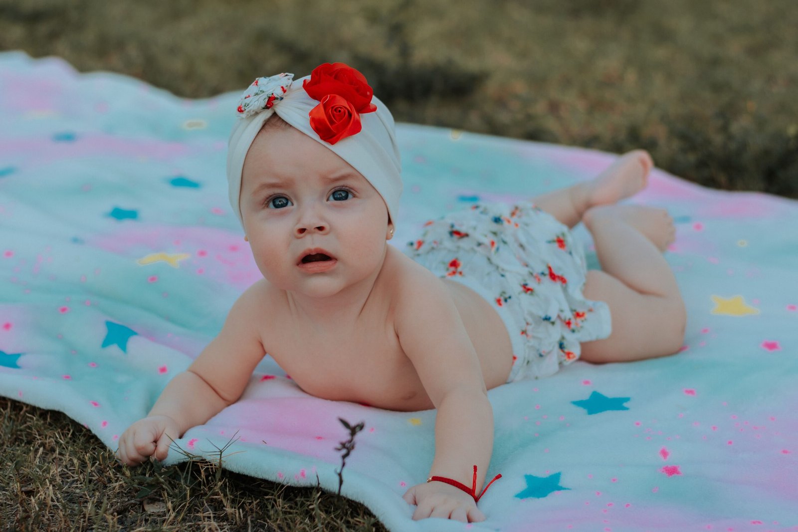 Adorable baby girl lying on a vibrant blanket outdoors with curious expression.