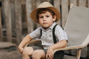 Adorable boy with straw hat seated on chair outdoors, exuding charm.