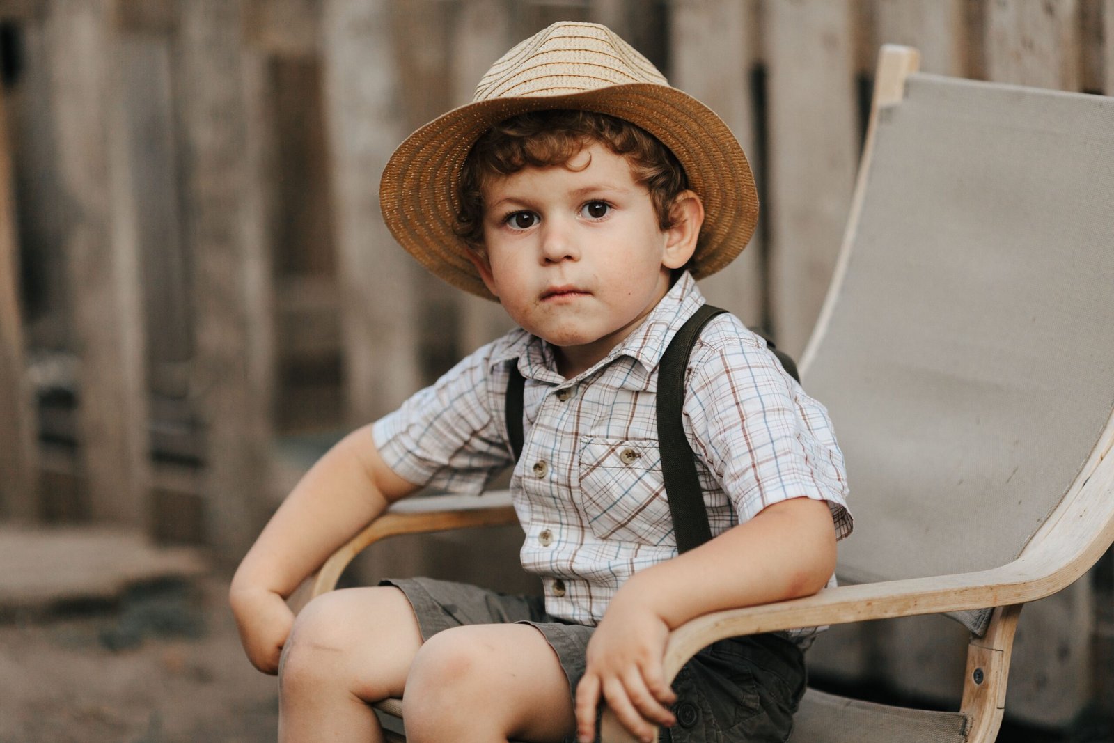 Adorable boy with straw hat seated on chair outdoors, exuding charm.